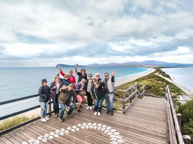 A group of tourists waving from a wooden lookout point over a sandy beach.